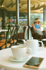 Cup of Hot Green Tea and White Teapot with a Mobile Phone on Cafe's Table  