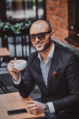 Elegant smiling man is drinking coffee while sitting outside at cafe on his break.