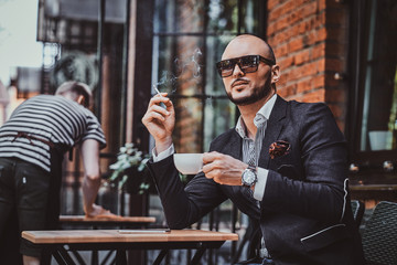 Pensive modern man is smoking a cigarette outside of coffeshop while drinking his cup of coffee.