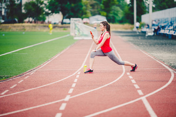 Sports fitness Caucasian women doing warm-up before exercise. Athlete stretching legs doing warm-up before training. Jogging in the city at the stadium. Girl uses headphones handset phone sportswear