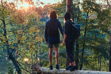 Romantic couple is standing on the log in green bright forest and watching sunset. They have backpacks and hats.