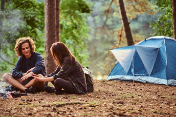 Young happy students are warming up near bonfire in the green forest. There are tent nearby.