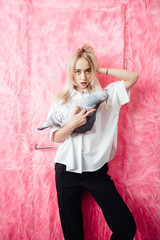 Young girl blogger dressed in stylish white shirt and black trousers poses with with a soft toy in the shape of a pigeon on the background of pink fur wall