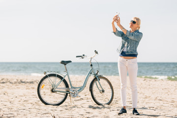 Obraz na płótnie Canvas Pretty blonde girl in white pants and denim coat taking selfie on the beach with bicycle. Atrractive woman relaxing near the sea after bike ride