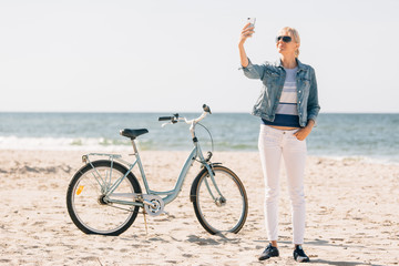 Pretty blonde girl in white pants and denim coat taking selfie on the beach with bicycle. Atrractive woman relaxing near the sea after bike ride