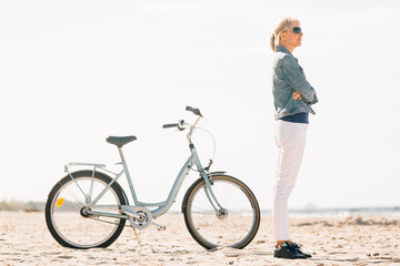 Pretty blonde girl in white pants and denim coat standing on the beach with bicycle. Atrractive woman relaxing near the sea after bike ride