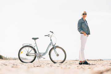 Fototapeta premium Pretty blonde girl in white pants and denim coat standing on the beach with bicycle. Atrractive woman relaxing near the sea after bike ride