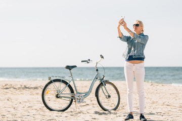 Naklejka premium Pretty blonde girl in white pants and denim coat taking selfie on the beach with bicycle. Atrractive woman relaxing near the sea after bike ride