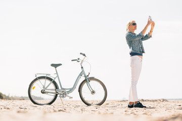Pretty blonde girl in white pants and denim coat taking selfie on the beach with bicycle. Atrractive woman relaxing near the sea after bike ride