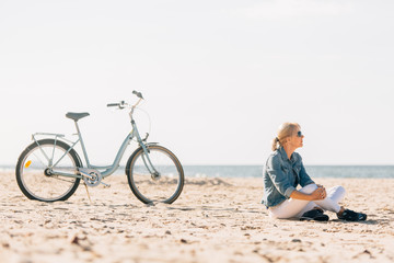 Pretty blonde girl in white pants and denim coat sitting on the beach near bicycle. Atrractive woman relaxing near the sea after bike ride