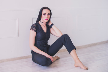 barefoot brunette model in a black suit sitting on the floor. Hand in hair. Against the background of a white wall.