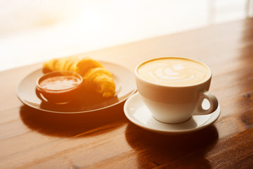 Cup of cappuccino with latte art and croissant with jam on the wooden table in cafe in the morning