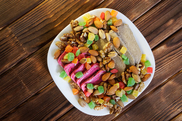 Arab sweets: unusual pink and traditional gray halvah in a white plate next to candied fruits, pistachios, almonds and walnuts. National Iranian dishes. Natural wooden background