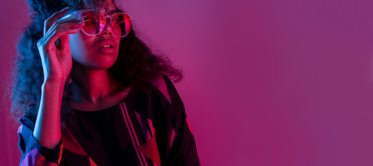 Mode junge afrikanische Mädchen schwarze Frau trägt eine stilvolle rosa Sonnenbrille mit Blick auf die Kamera einzeln auf lila Studiohintergrund der Party, horizontales Banner für Website-Design, Porträt, Kopienraum