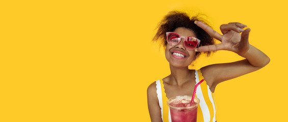 Happy young african woman hold drink wear glasses laugh look at camera, cheerful black teen girl enjoy summer cocktail isolated on yellow studio background, banner design close up portrait copy space
