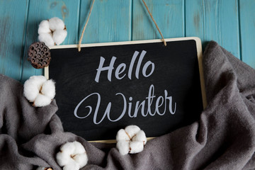 Hello winter postcard. Winter banner with warm gray shawl, cotton flowers on a blue wooden background