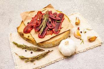 Basturma, dried tenderloin of beef meat, jerky, thinly sliced. Dry rosemary, pepper mix, chili, garlic on vintage linen cloth. Delicious food on a wooden board, stone background, close up.