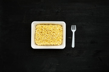 Instant noodles in container with plastic fork on a black wooden table, top view