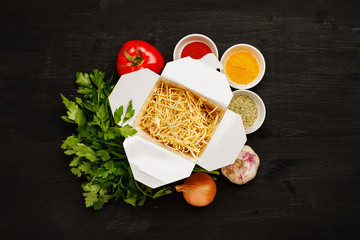 Dry noodles in paper box with greenery, vegetables and spices on a black table, top view