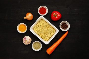 Instant noodles in contaiber with vegetables and spices on a black wooden table, top view