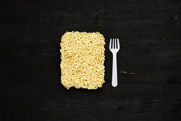 Briquet of instant noodles with plastic fork on a black wooden table, top view