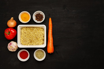 Instant noodles in contaiber with vegetables and spices on a black wooden table, top view, text space