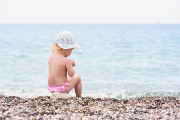 Cute toddler girl sits on the sea beach in hot summer day and plays with stones.