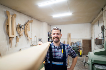 Portrait of smiling bearded carpenter woodworker holding wooden plank on shoulder ready to do his next project in carpentry workshop.