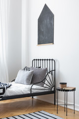 Cushions on bed next to table in black and white bedroom interior with blackboard on the wall and small metal bedside table, real photo with mockup