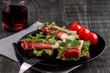 On the table there is a dish with jamon sandwiches, tomato, greens and a glass of red wine