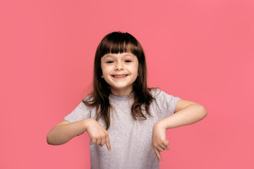 Portrait of a beautiful young little girl child wearing grey t-shirt standing isolated over pink background, presenting copy space down here and smiling. Positive and happy expression