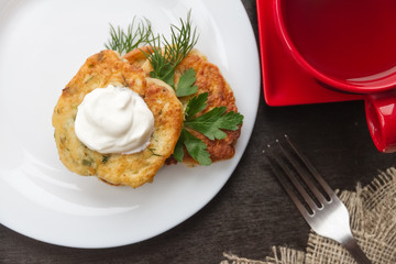 cheesecakes with sour cream and green seasoning on a white plate, next to red cup and metal fork in sackcloth