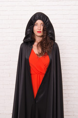 Naklejka premium Her sorcery skills are just far too impressive. Halloween party girl. Sexy girl wearing hooded black cloak on white brickwall. Mysterious girl with classic witch look. A girl changed into a witch