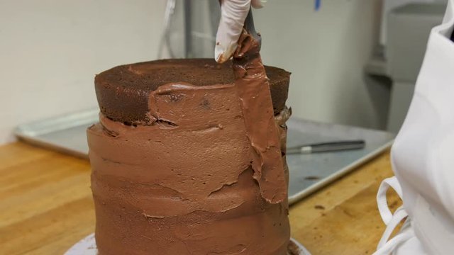 Baker Adds Final Layer To Tired Chocolate Cake. Baker frosts the top layer of a tiered, round chocolate cake with a flat spatula. The frosting is spread evenly around the whole cake.