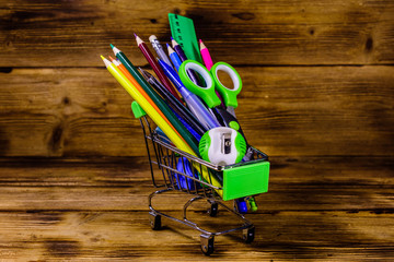 Small shopping cart with different school stationery on a wooden background. Back to school concept
