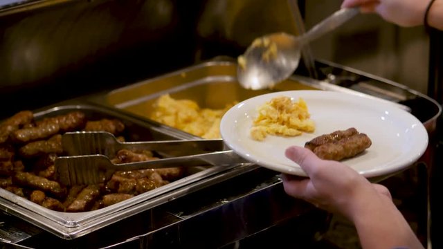 Woman Dishes Up Breakfast From Buffet Line This over the shoulder shot shows a woman dishing scrambled eggs onto her white plate with sausages in this hot breakfast buffet line.