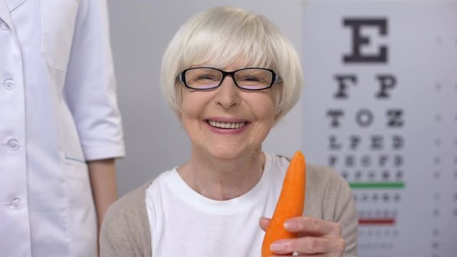 Oculist proposing carrot to smiling elderly lady, natural vitamins for sight