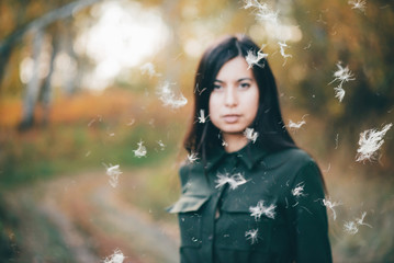 Dreamy beautiful girl in airy white fluff on bokeh background with yellow leaves. Inspired girl in autumn forest with golden foliage in blur. Blurred beauty portrait among flying spores of thistle.