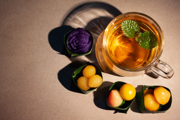 Thai sweets in banana leaves that are arranged with cup of tea on brown paper. Purple rose steamed layer cake, golden jackfruit seeds, thai mung bean marzipan and golden drops.