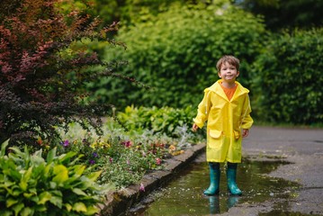 Little boy playing in rainy summer park. Child with umbrella, waterproof coat and boots jumping in...