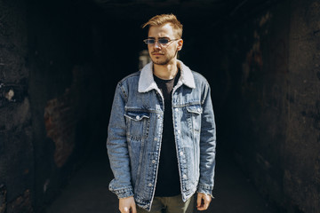 Portrait of young man in blue jeans jacket with sunglasses outdoor on dark background.