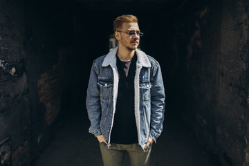 Portrait of young man in blue jeans jacket with sunglasses outdoor on dark background.