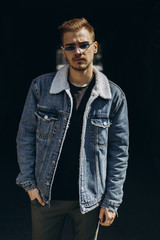 Portrait of young man in blue jeans jacket with sunglasses outdoor on dark background.