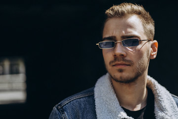 Portrait of young man in blue jeans jacket with sunglasses outdoor on dark background.
