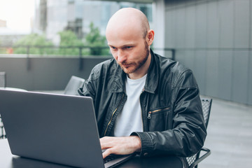 Attractive adult successful bald bearded man in black jacket with laptop in street cafe at city