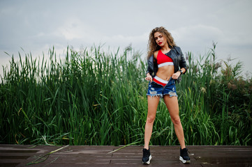 Sexy curly model girl in red top, jeans denim shorts, leather jacket and sneakers posed in the reed.