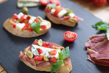 Traditional Italian bruschetta with blue cheese, feta, tomatoes, basil leaves, jamon on black stone background.