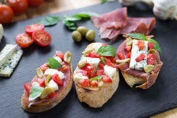 Traditional Italian bruschetta with blue cheese, feta, tomatoes, basil leaves, jamon on black stone background.