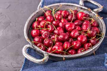 ripe, juicy cherries in round metal bowl with handles, on napkin color of jeans and on gray cement table