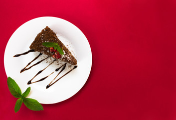 Chocolate cake slice with berries and leaves of mint on a white plate, top view on a red background. Copy space